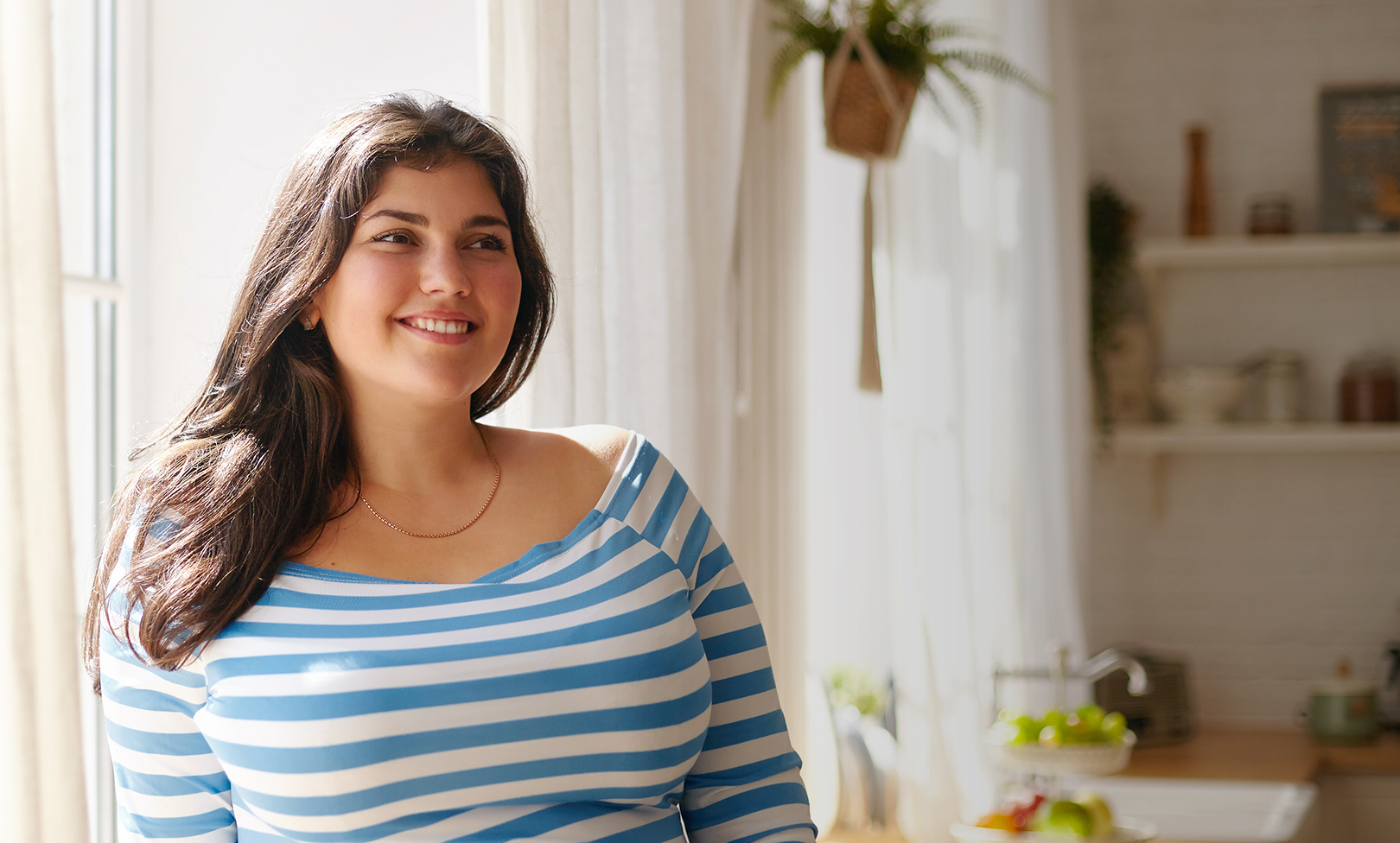 Beauty, bodypositivity, people and lifestyle concept. Indoor shot of gorgeous brunette Latin girl posing by window in kitchen dressed in xxl blue and white striped shirt, having joyful beaming smile