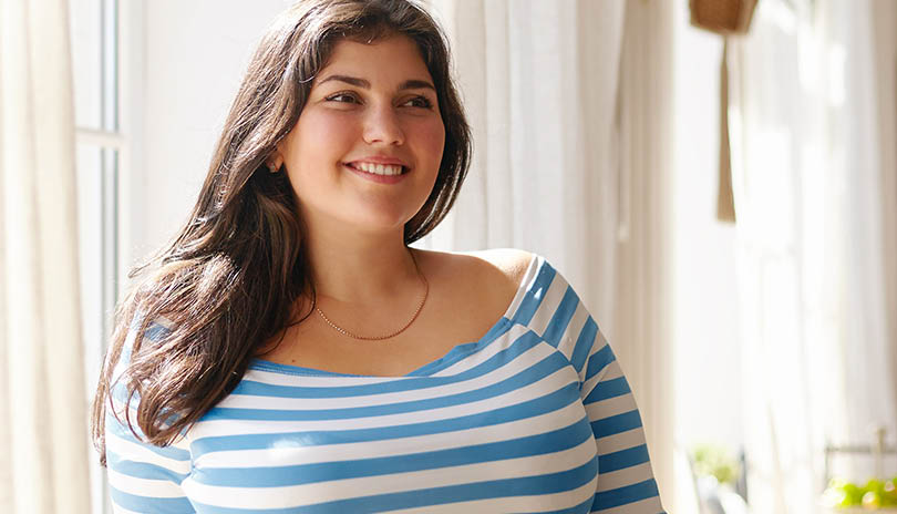 Beauty, bodypositivity, people and lifestyle concept. Indoor shot of gorgeous brunette Latin girl posing by window in kitchen dressed in xxl blue and white striped shirt, having joyful beaming smile